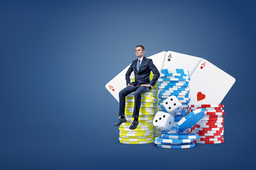 A tiny businessman sits on a stack of casino chips near large dice and playing cards.