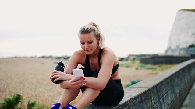 Young sporty woman with smartphone sitting outside, texting.