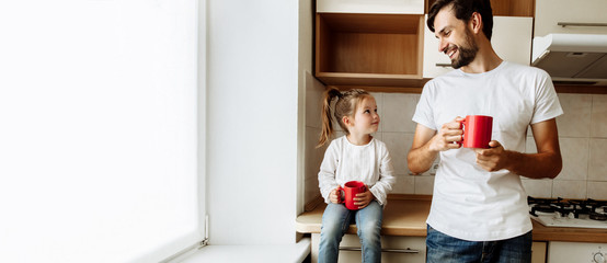 Parenthood. Family. Love. Dad and his little daughter are holding red cups, looking at each other and smiling. In the kitchen at home