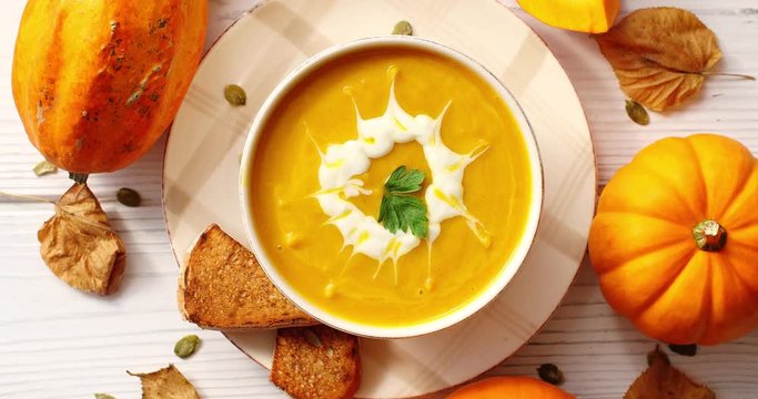 From above view of creamy yellow pumpkin soup in bowl with bread and pumpkins placed around on wooden background