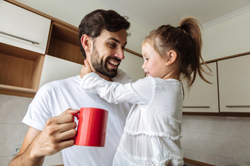 Parenthood. Family. Home. Dad is holding a cup and his little daughter in his arms. Both are talking and smiling. In the kitchen at home