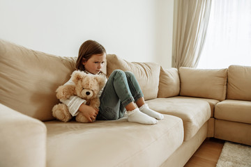 Childhood. Education. Problem. Offended little girl is hugging her teddy bear while sitting alone on a couch at home