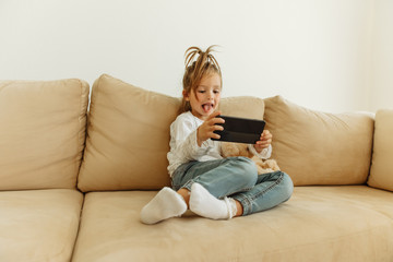 Childhood. Technologies. Fun. Little girl is using a smartphone and showing her tongue while sitting on a couch at home