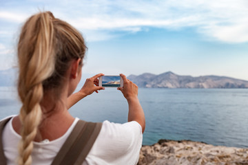 Hiker girl with smartphone taking landscape photo of sea bay while traveling