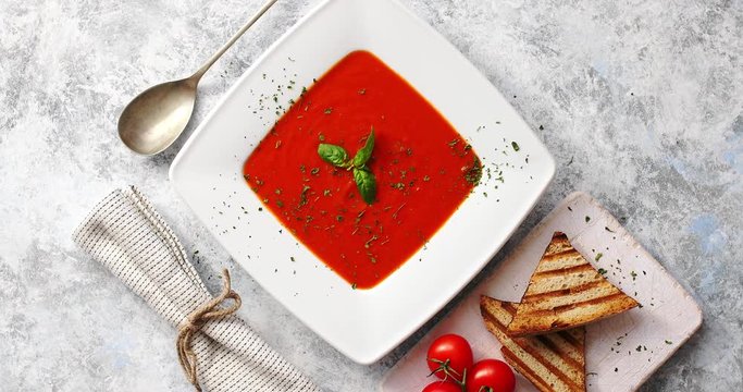 From above view of red tomato soup in white plate served with crisp bread and tablespoon on gray background 
