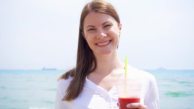 Smiling vegan woman in white shirt holding cup with strawberry smoothie against sea in slow motion. Fit vegetarian female enjoying healthy lifestyle outdoors
