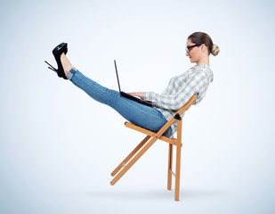 Young girl with glasses balances on a wooden chair working on a laptop, concept of relaxation at...