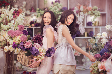 Beautiful asian florist girls holding bouquet and basket of flowers for sale against floral bokeh background in flower shop indoors. Two attractive asian females florists working in retail store. 2