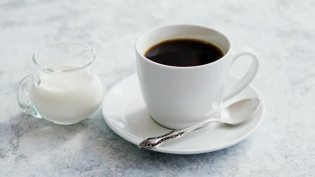 Arrangement of small glass pitcher with fresh milk on marble table with white cup of coffee with silver spoon on saucer