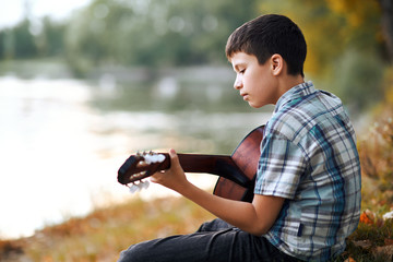 the boy plays an acoustic guitar, sits on the Bank of the river, autumn forest at sunset, beautiful nature and the reflection of trees in the water