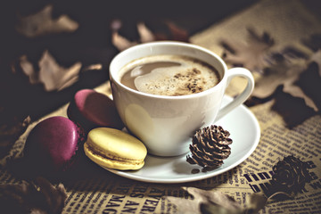 Autumn, fall dried leaves, hot steaming cup of cappuccino coffees, macaroons, cones, newspaper on wooden table background. Seasonal, evening coffee, Sunday relaxing and still life concept. Flat lay. 