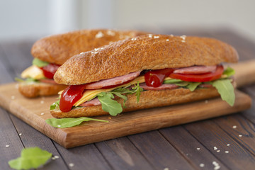  Two baguette sandwiches with salami, cheese, arugula salad, tomatoes and onion on a cutting board. Sandwiches on dark wooden background. Tasty snack.