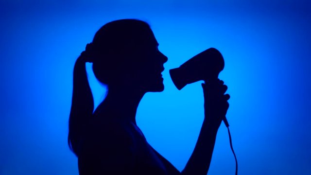 Silhouette of young woman having fun singing into hair dryer. Female's face in profile pretending to hear music dancing silly on blue background. Black contour shadow of teenager's half-face singing
