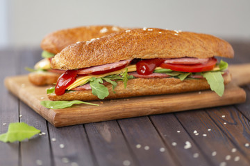  Two baguette sandwiches with salami, cheese, arugula salad, tomatoes and onion on a cutting board. Sandwiches on dark wooden background. Tasty snack.
