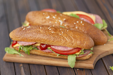  Two baguette sandwiches with salami, cheese, arugula salad, tomatoes and onion on a cutting board. Sandwiches on dark wooden background. Tasty snack.