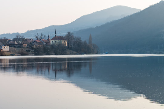 Small church on Elbe riverside with blue hills in background. Reflection of the scenic landscape in calm water. Spring, Czech Republic, Northern Bohemia.