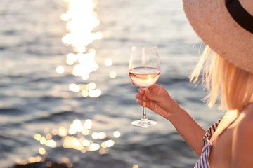  Girl holds wineglass with white wine in hand on beach at summer background of sunset sea or ocean. Blond beautiful woman is straw hat is relaxing, drinking, traveling and enjoying life in vacation. © Marina April