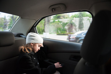 sad lonely small girl in wool hat and coat sitting inside car near window