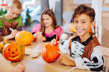 Fototapeta premium Amazing day. Dark-eyed cute and funny boy wearing skeleton costume for Halloween feeling amazing coloring pumpkins