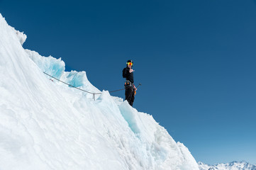 Naklejka na ściany i meble A mountaineer stands on the edge of a glacier with a snow shovel in his hands and shows Shak's gesture against the blue sky
