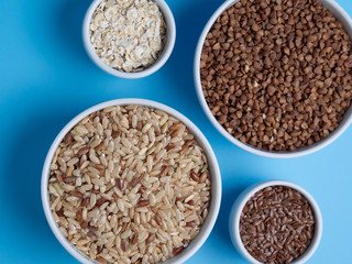 brown rice, buckwheat, flax and oat flakes in white cups on a blue background