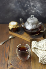 Tea in glass cup with teapot and knitted blanket near, with jam in jar at wood background, with spoon near.
