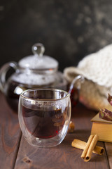 Tea in glass cup with teapot and knitted blanket near, with jam in jar at wood background, with spoon near.