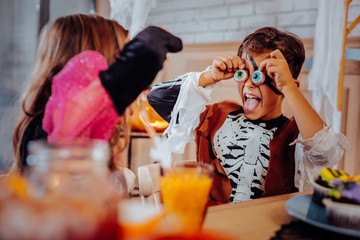 Eyes to face. Cute dark-haired boy putting sweet cookies in the form of eyes to his face celebrating Halloween