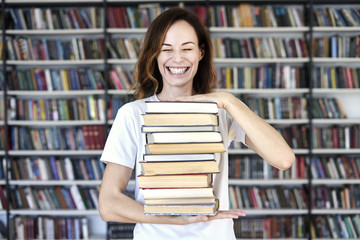 Woman model college student with books at library holds bunch of books, laughs looks smart, smiling to camera.