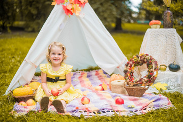 Little girl lying and playing in a tent, children's house wigwam in park Autumn portrait of cute curly girl. Harvest or Thanksgiving. autumn decor, party, picnic. Child in yellow dress with apple  © MartaKlos