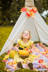 Little girl lying and playing in a tent, children's house wigwam in park Autumn portrait of cute curly girl. Harvest or Thanksgiving. autumn decor, party, picnic. Child in yellow dress with apple  © MartaKlos