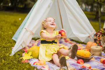 Little girl lying and playing in a tent, children's house wigwam in park Autumn portrait of cute curly girl. Harvest or Thanksgiving. autumn decor, party, picnic. Child in yellow dress with apple  © MartaKlos