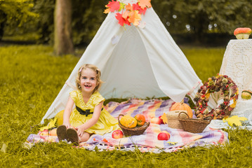 Fototapeta na wymiar Little girl lying and playing in a tent, children's house wigwam in park Autumn portrait of cute curly girl. Harvest or Thanksgiving. autumn decor, party, picnic. Child in yellow dress with apple 