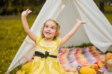 Little girl lying and playing in a tent, children's house wigwam in park Autumn portrait of cute curly girl. Harvest or Thanksgiving. autumn decor, party, picnic. Child in yellow dress with apple  © MartaKlos