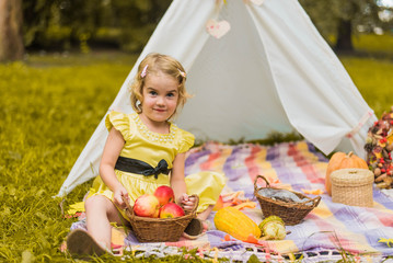 Little girl lying and playing in a tent, children's house wigwam in park Autumn portrait of cute curly girl. Harvest or Thanksgiving. autumn decor, party, picnic. Child in yellow dress with apple  © MartaKlos
