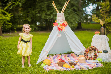 Little girl lying and playing in a tent, children's house wigwam in park Autumn portrait of cute curly girl. Harvest or Thanksgiving. autumn decor, party, picnic. Child in yellow dress with apple  © MartaKlos