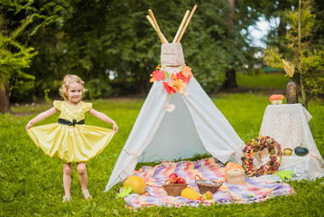 Little girl lying and playing in a tent, children's house wigwam in park Autumn portrait of cute curly girl. Harvest or Thanksgiving. autumn decor, party, picnic. Child in yellow dress with apple  © MartaKlos