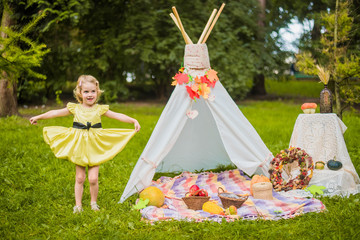 Little girl lying and playing in a tent, children's house wigwam in park Autumn portrait of cute curly girl. Harvest or Thanksgiving. autumn decor, party, picnic. Child in yellow dress with apple  © MartaKlos