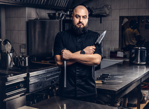 Bearded cook in black uniform holds knife and standing with crossed arms in the kitchen.