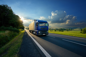 Fototapeta na wymiar Blue truck arriving on the asphalt road in rural landscape in the rays of the sunset