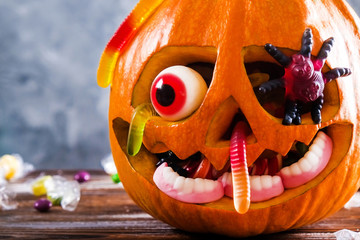 Orange pumpkin carved into Jack o Lantern with eyeball candy and gummy worms & spiders on wooden...