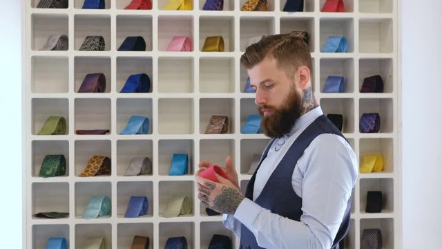 Tattooed bearded male dressed in elegant suit chooses his tie near a rack in a menswear store.