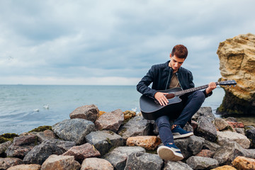 Young man with acoustic guitar playing on beach surrounded with rocks on rainy day