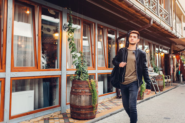 Young man with acoustic guitar in case walking on city street by cafe. Hipster college guy carrying...