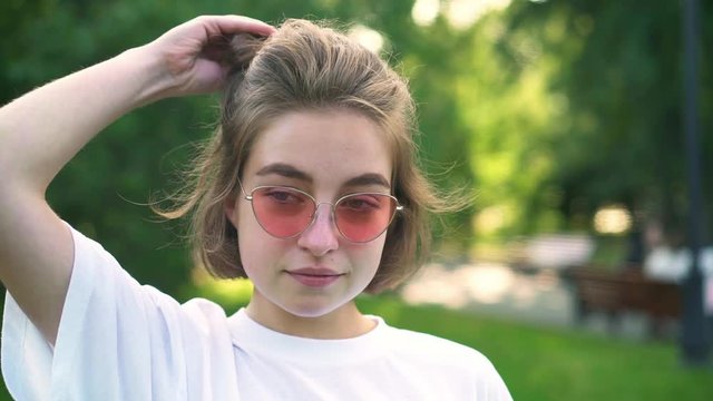 Attractive young woman in pink sunglasses and white t shirt taking off her rubber band and playing with her hair in summer park. Concept of fashion and accessories. Slider slow motion portrait shot