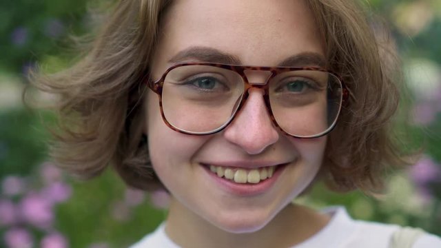 Young woman with short fair hair in white t shirt puts on her glasses and smiles cheerfully looking at camera. Facial expressions and emotions concept. Handheld real time close up shot