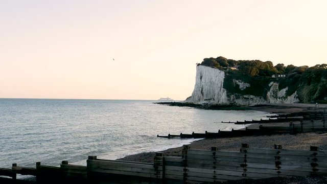 Waves wash ashore at the shadowy white cliffs of Dover in the evening as a ferry boat in the far distant background departs from the ferry port of Dover on the English Channel