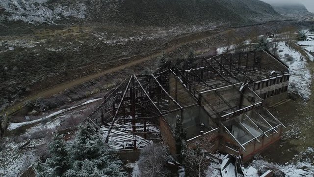 Aerial drone scene of destroyed building, old burned hotel in snowy grey day in Tunuyan river Valley. Camera moves to senital view. Naked metal roof structure. Snowy mountains, vegetation 