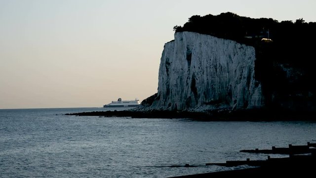 Waves roll gently ashore with the iconic white cliffs of Dover in the evening shadows with a ferry boat passing behind the cliffs inbound for the ferry port at Dover.