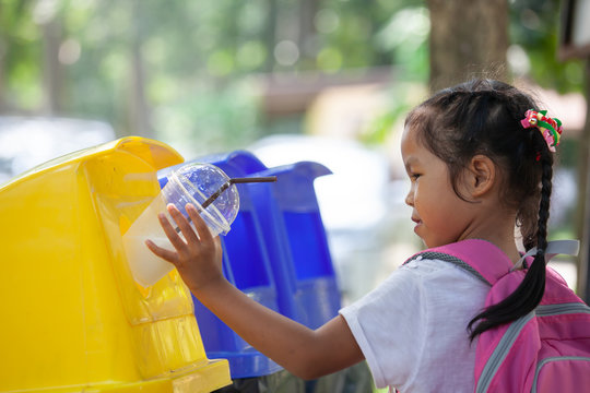 Cute asian child girl throwing plastic glass in recycling trash bin at public park
